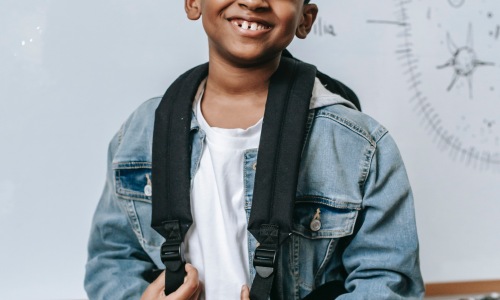 Kid smiling at the camera while standing in classroom with backpack on